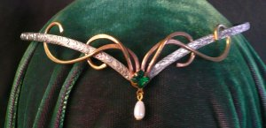 An example of an aurora circlet, one of our many handmade circlets with brass swirl flourishes soldered to a silver plated band, with a small green stone  in gold tone setting soldered to the front and a faux pearl dangle.