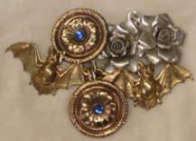 Several cloak clasps in a variety of metal finishes including silver, gold and brass including some mass produced and some handmade in zoomorphic shapes such as bats, floral designs such as roses and a round pair with blue stones.