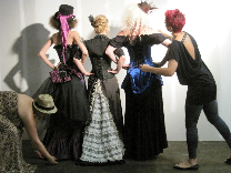 A group of models dressed for a steampunk themed photoshoot wearing corsets, bustles, overskirts, modern Victorian skirts, hats, goggles and jewelery in black, magenta, blue and white.