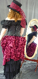 A woman dressed in a steampunk outfit, which consists of a mad hatter top hat with lace decoration, black corset over black peasant blouse and a red crushed velvet tie-front overskirt which can also serve as a bustle.